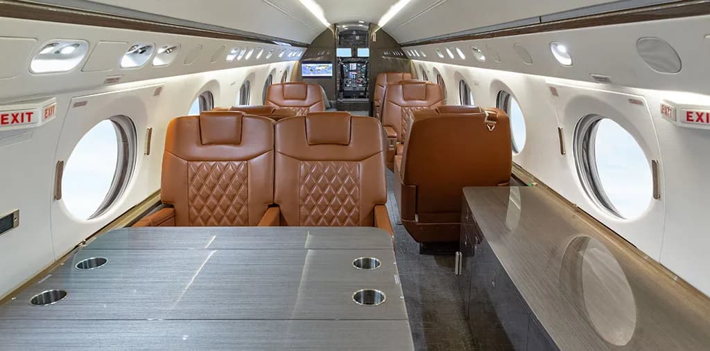 The spacious interior of a private jet cabin with brown leather seating and a table, looking towards the cockpit.