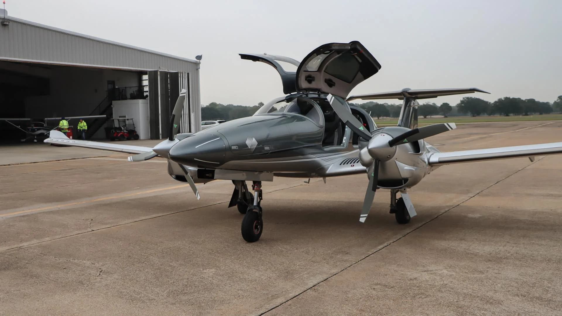A front-side view of the gray and silver Diamond DA62 aircraft with its gull-wing doors open, parked on an airport tarmac.