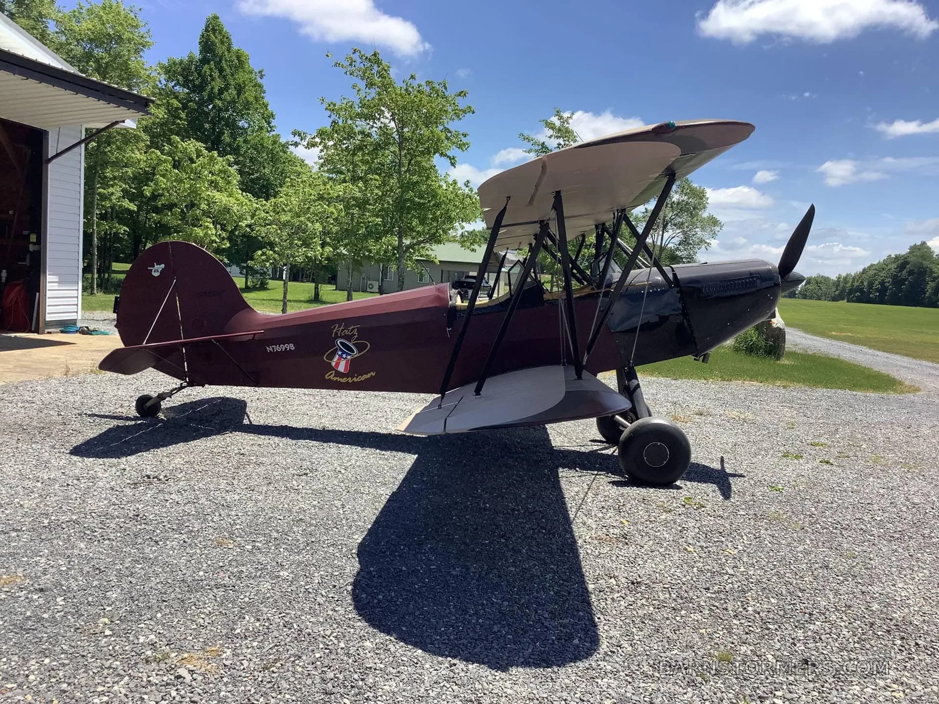 A maroon and black biplane with a 'Hotz American' logo on the fuselage, parked on a gravel path next to a building.