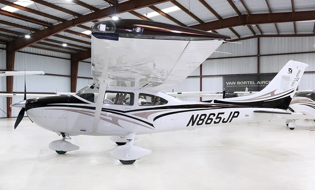 A white and black Cessna Skylane N865JP with brown accents is parked in a hangar, viewed from the front-right side.