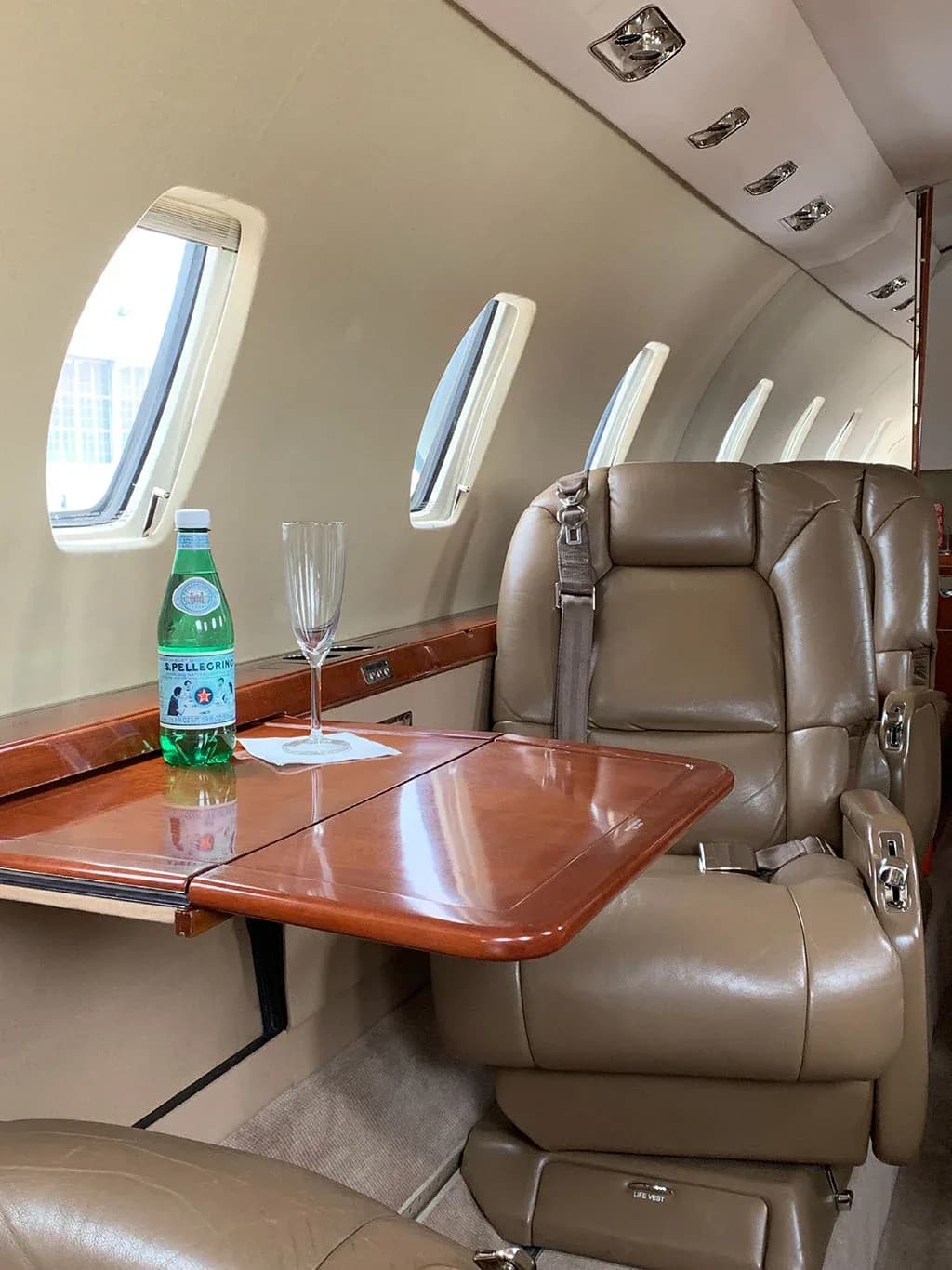 Close-up of a tan leather seat and a fold-out table with a bottle of sparkling water and a glass in the aircraft cabin.