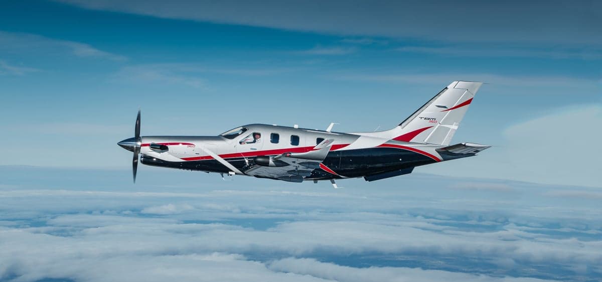A TBM 960 aircraft with a silver, black, and red paint scheme flying above clouds under a blue sky.