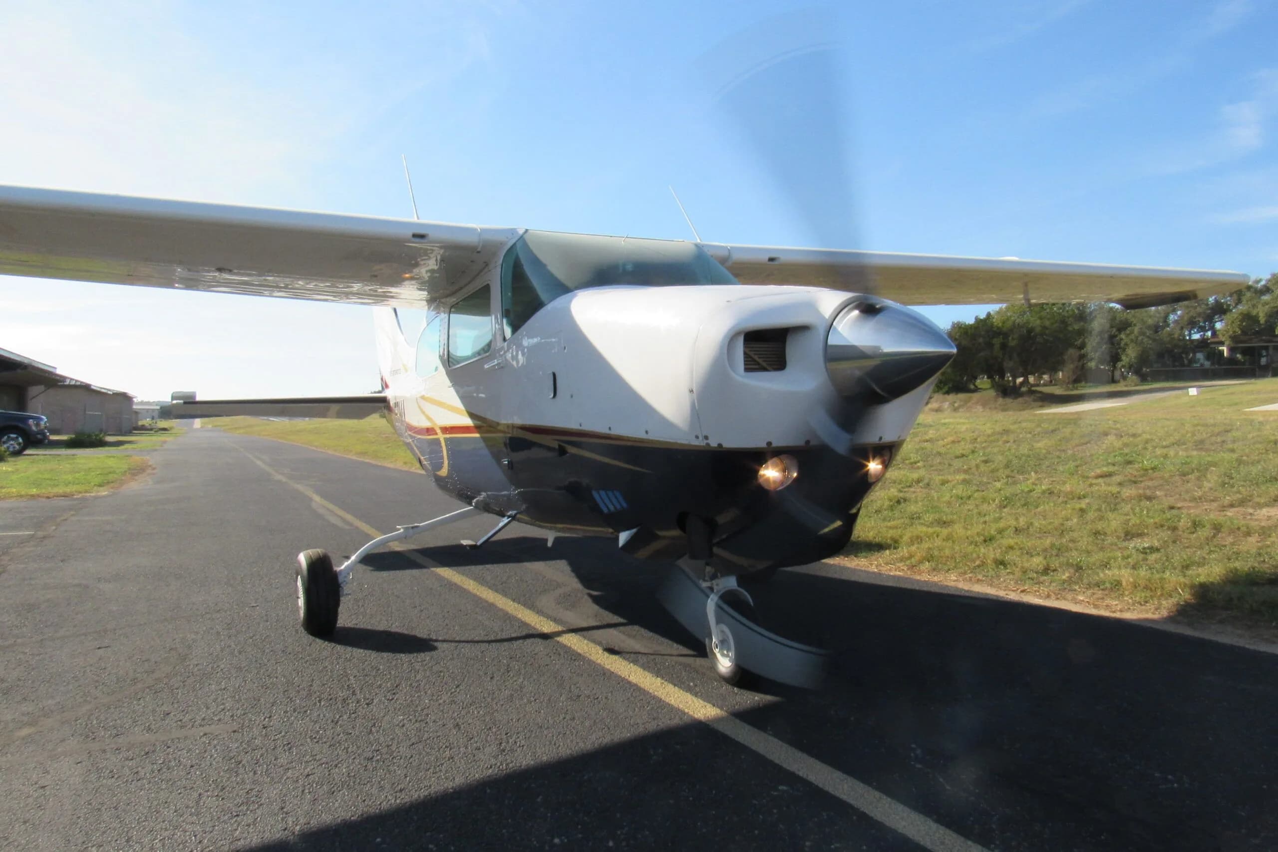 A front view of the white and blue Cessna 210, N58MA, with its propeller spinning, on an asphalt taxiway.
