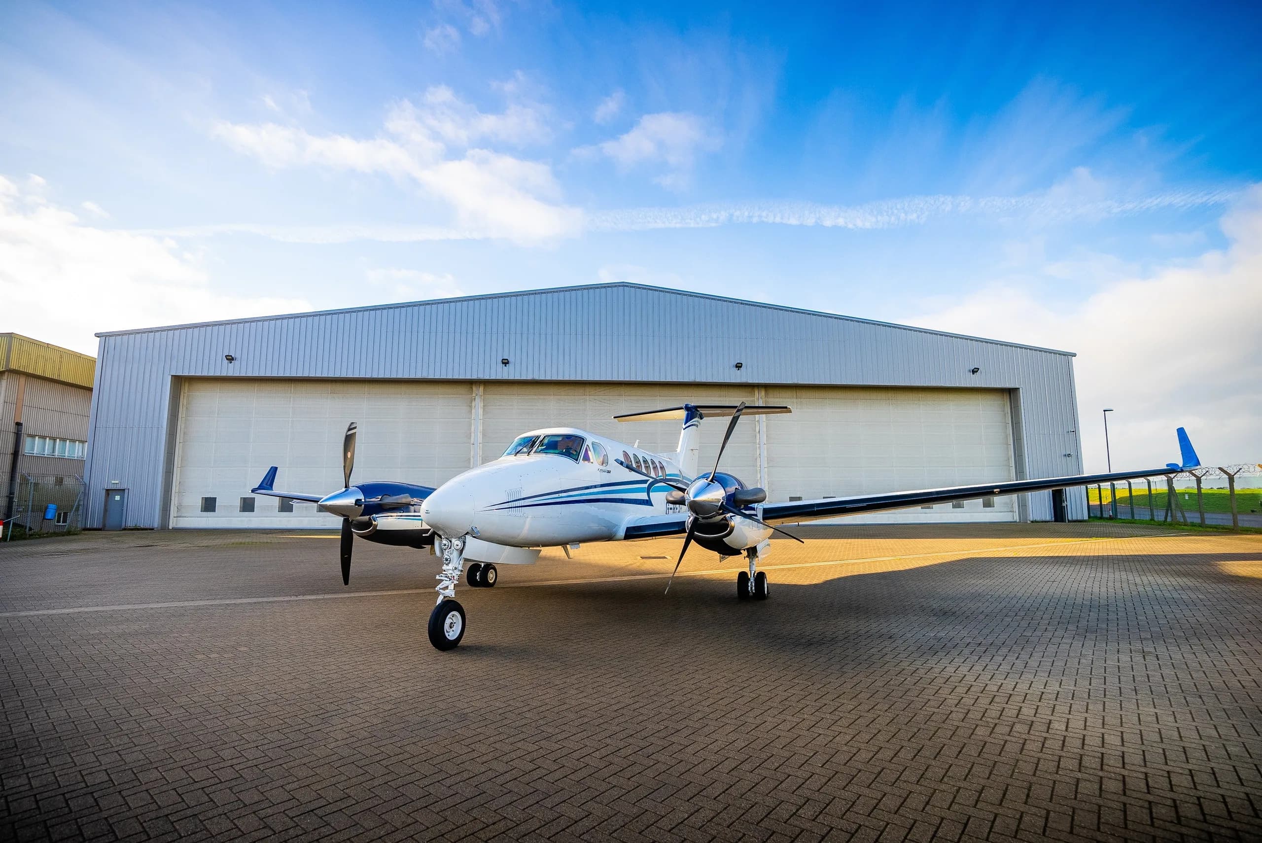 A white and blue Beechcraft King Air 260 parked on a tarmac in front of a hangar under a blue sky.