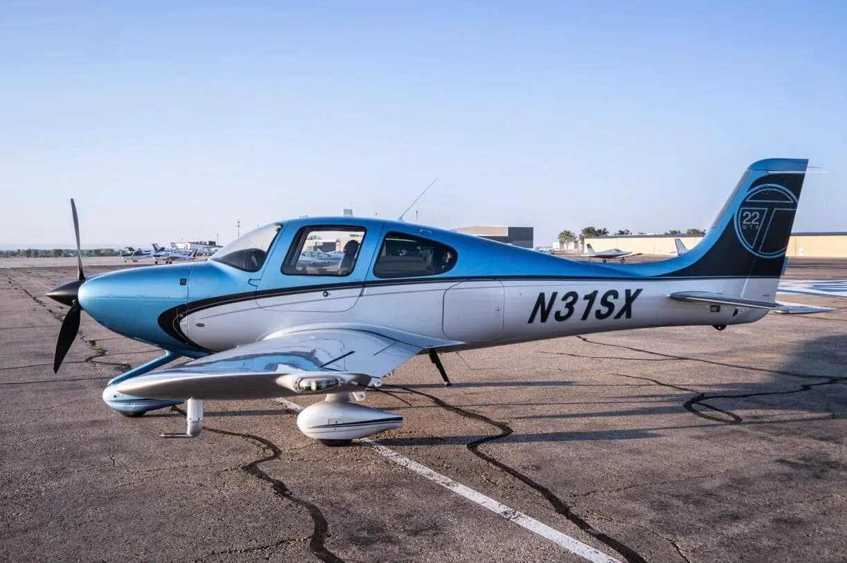 A Cirrus SR22T aircraft, N31SX, in blue and silver paint, parked on an airfield with other planes in the distance.