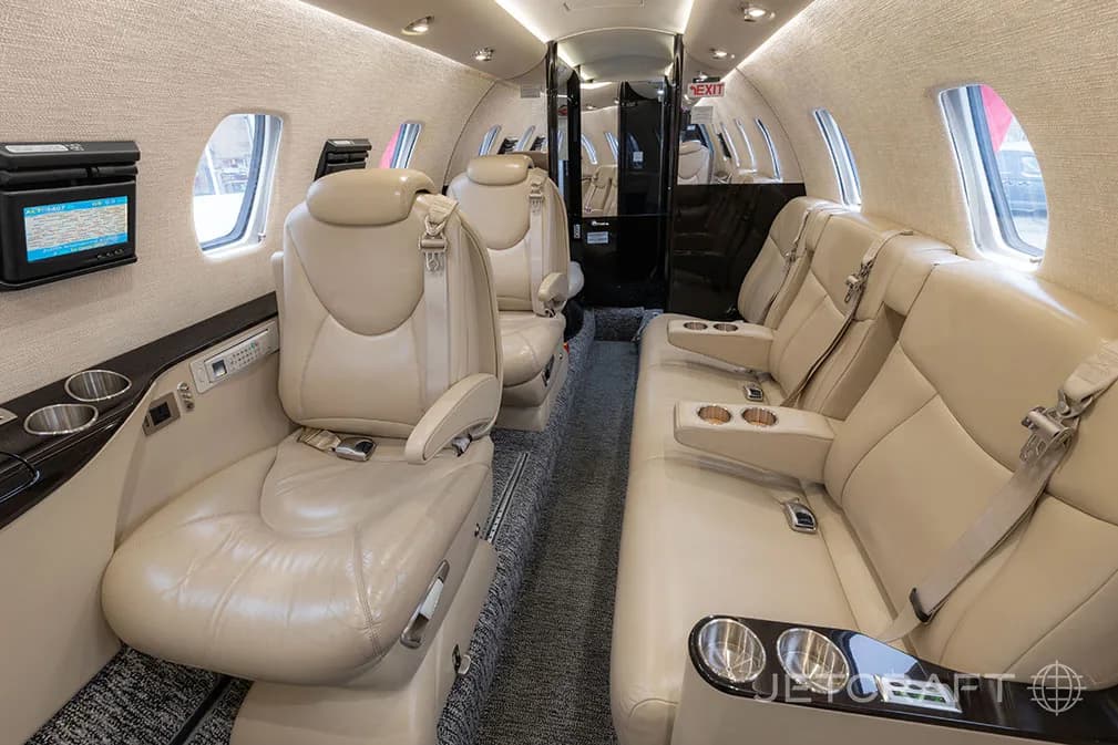 A view of the business jet's interior cabin, featuring light beige leather seating, fold-out tables, and cup holders.