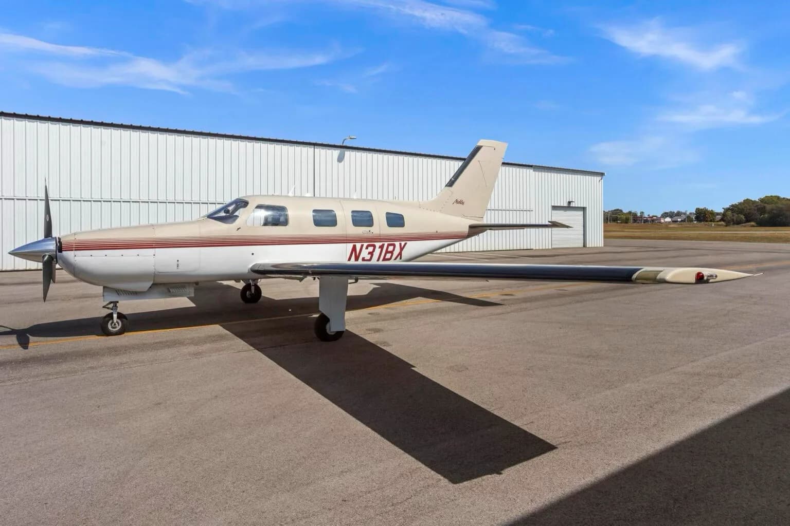 A Piper Malibu aircraft, N31BX, in white and tan with red stripes, parked on an airfield next to a hangar.