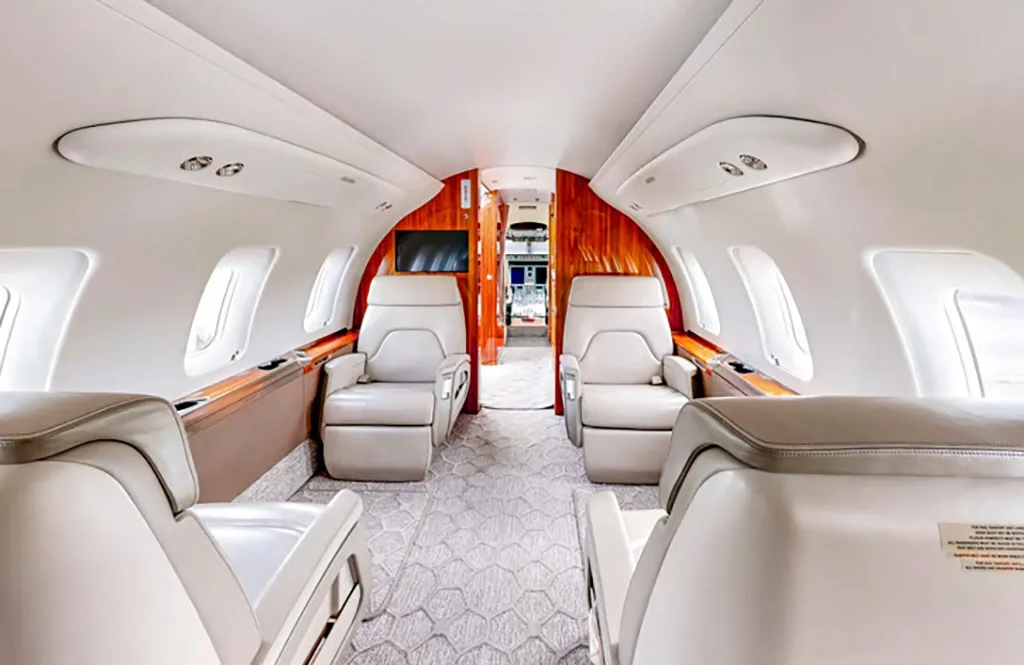 The spacious interior of a private jet cabin with light-colored seating and wood accents.