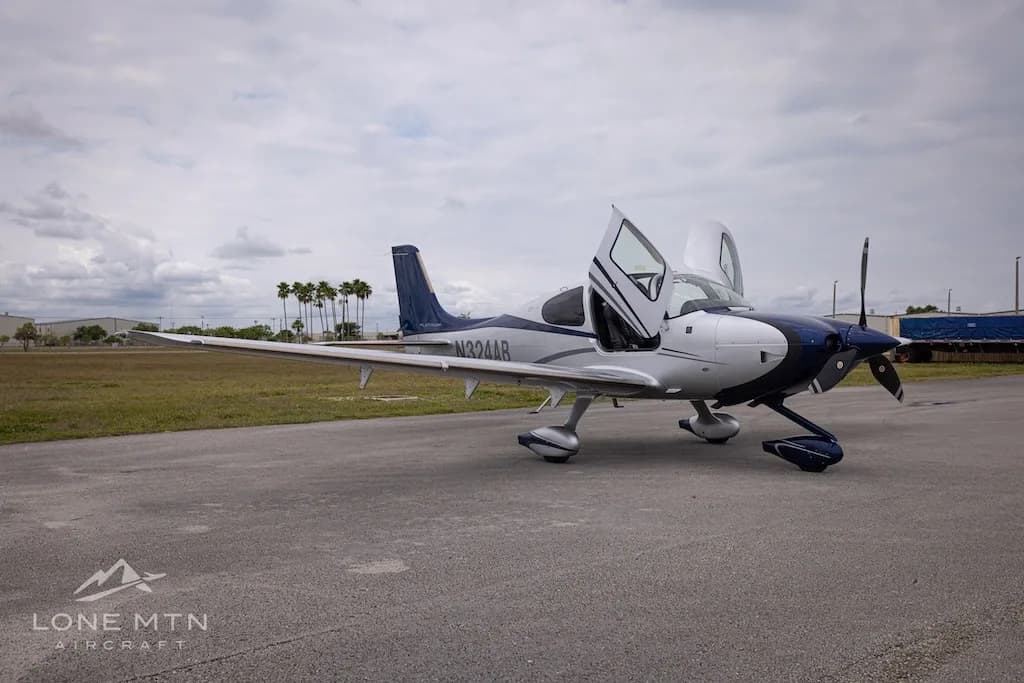 A white and blue Cirrus SR22T aircraft with open doors parked on an airfield, viewed from the front-left.