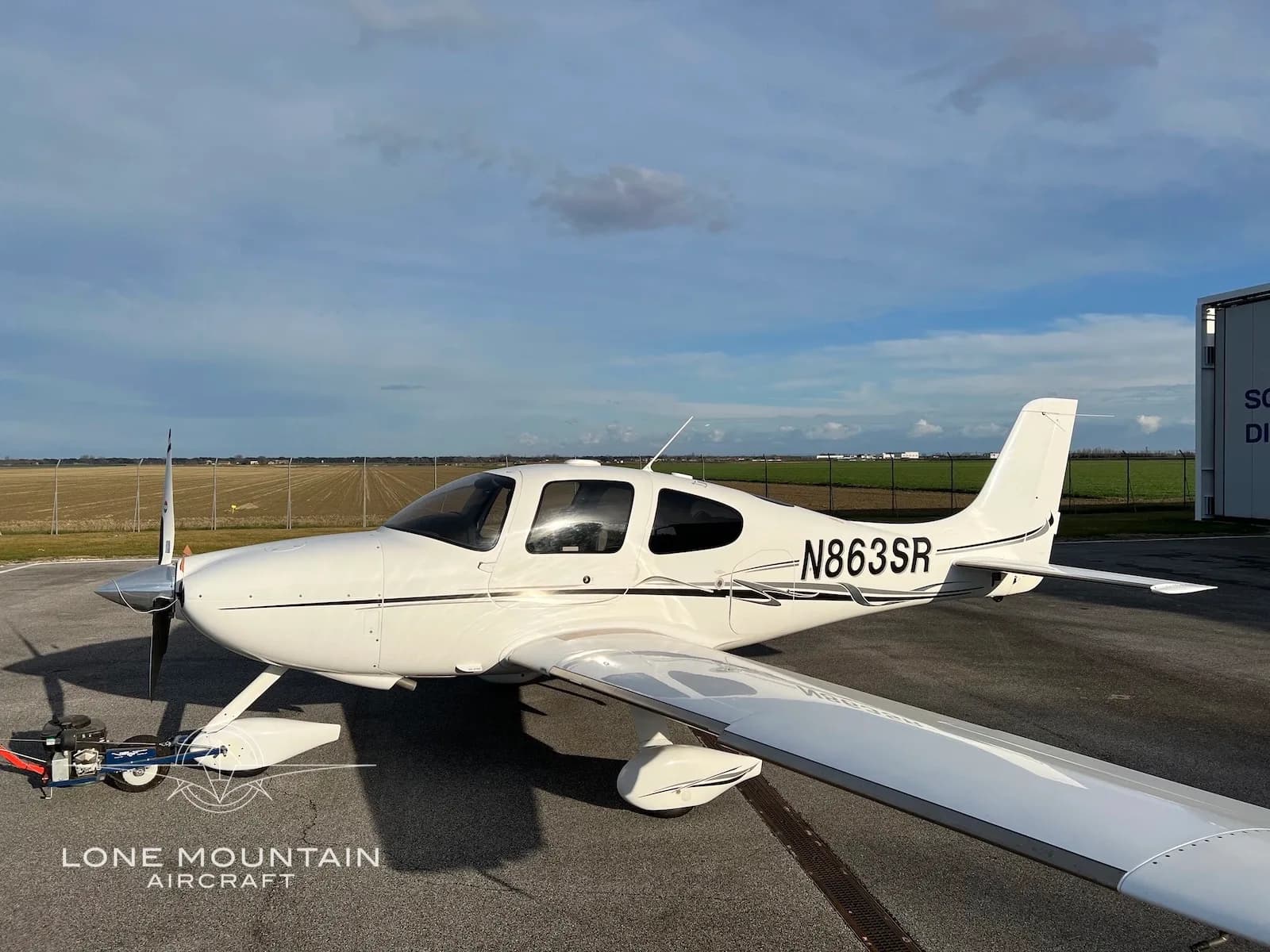A white Cirrus SR22 aircraft with registration N863SR is parked on an airfield under a partly cloudy sky, with a hangar in the background.