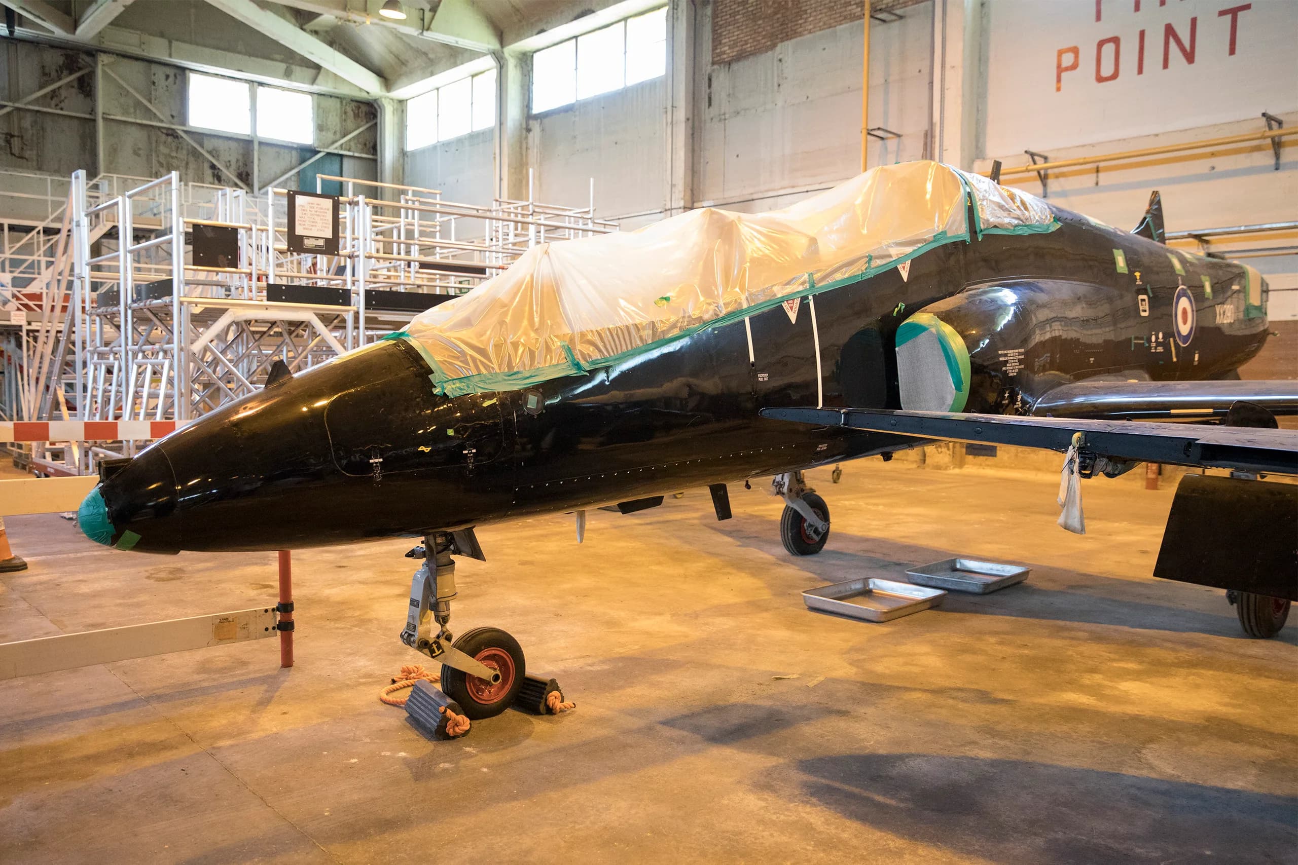 A black jet aircraft, possibly a BAE Hawk, partially covered with plastic sheeting in a hangar.