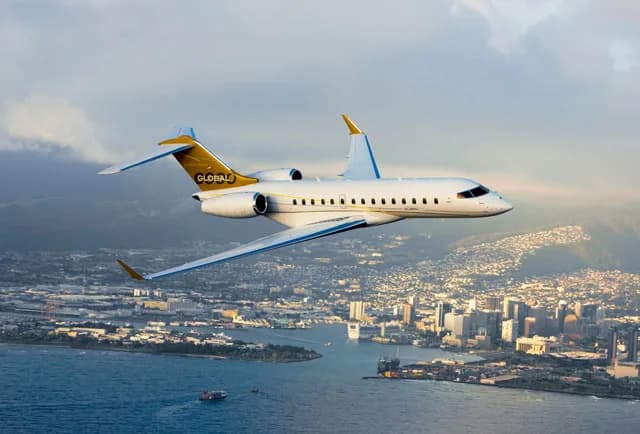 A white and gold Bombardier Global business jet flies over a coastal city with mountains in the background.
