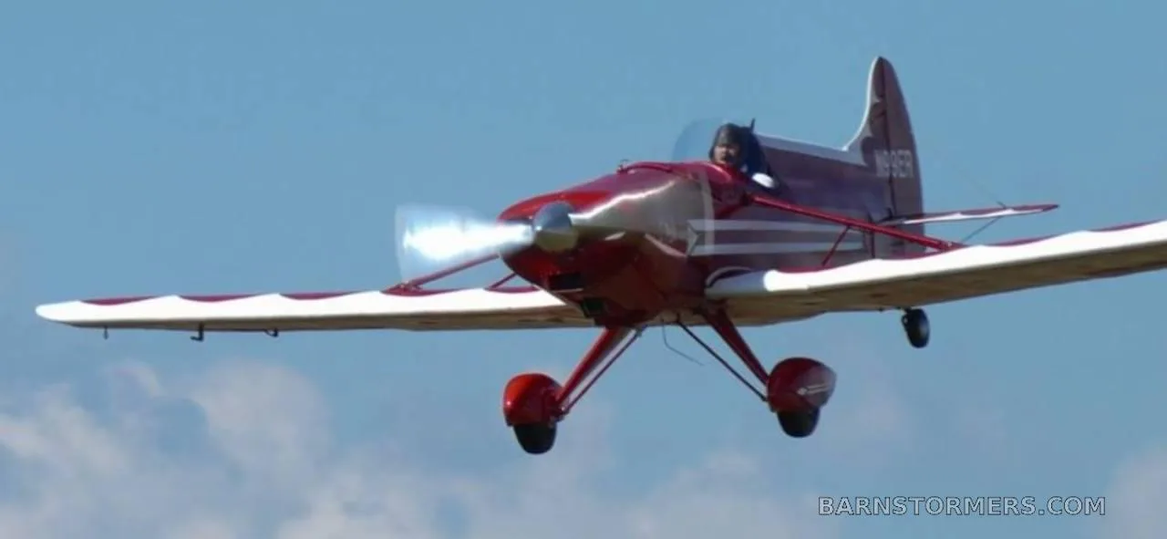 A red and white Spezio Tuholer monoplane, registration N99ER, with a pilot visible, flying against a clear blue sky.