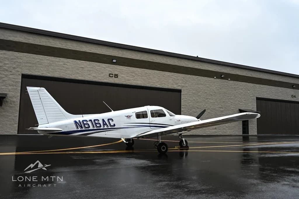A white single-engine aircraft, N616AC, with blue stripes is parked on a wet tarmac in front of hangars.