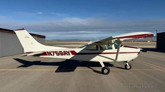 A white Cessna 172 with red and maroon stripes, registration N759AY, parked on an asphalt ramp next to a hangar.