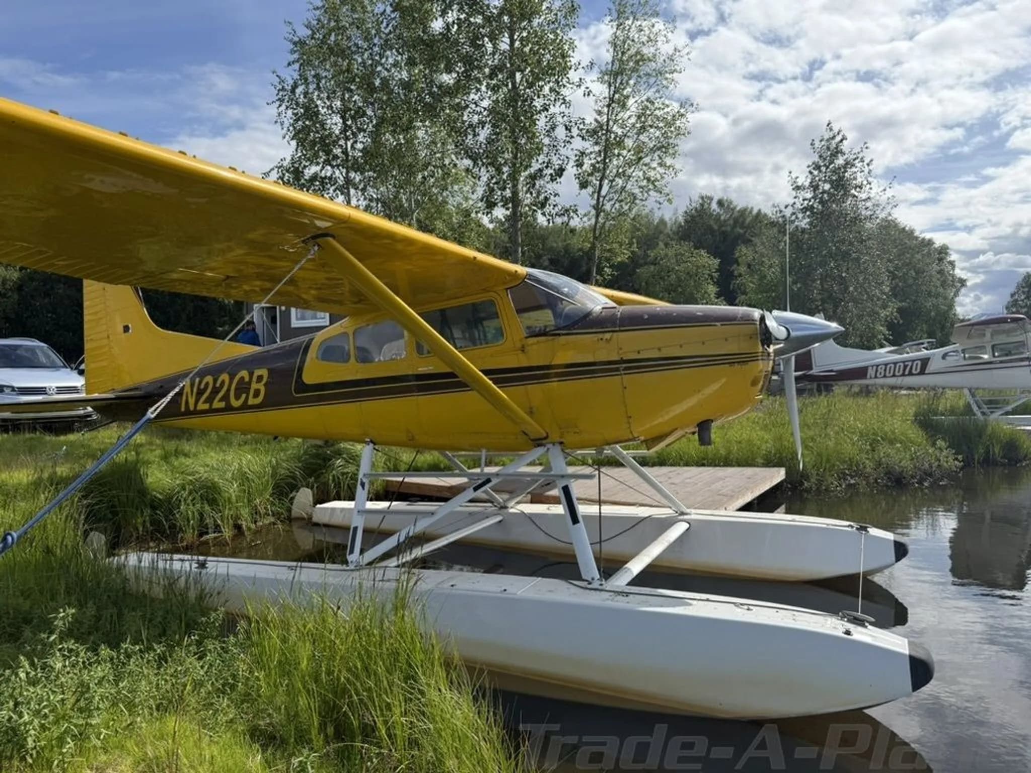 A yellow and brown floatplane, N22CB, is tied to a wooden dock on a grassy shore.