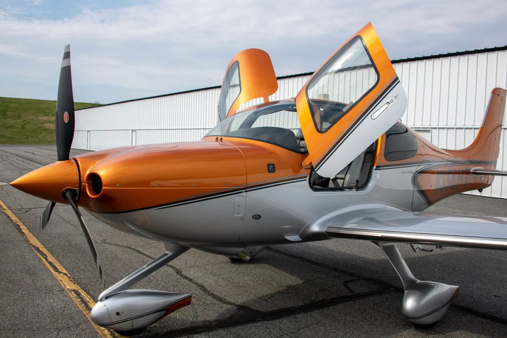 A front-side view of a metallic orange and silver Cirrus SR22 aircraft (N356HT) with its gull-wing doors open.