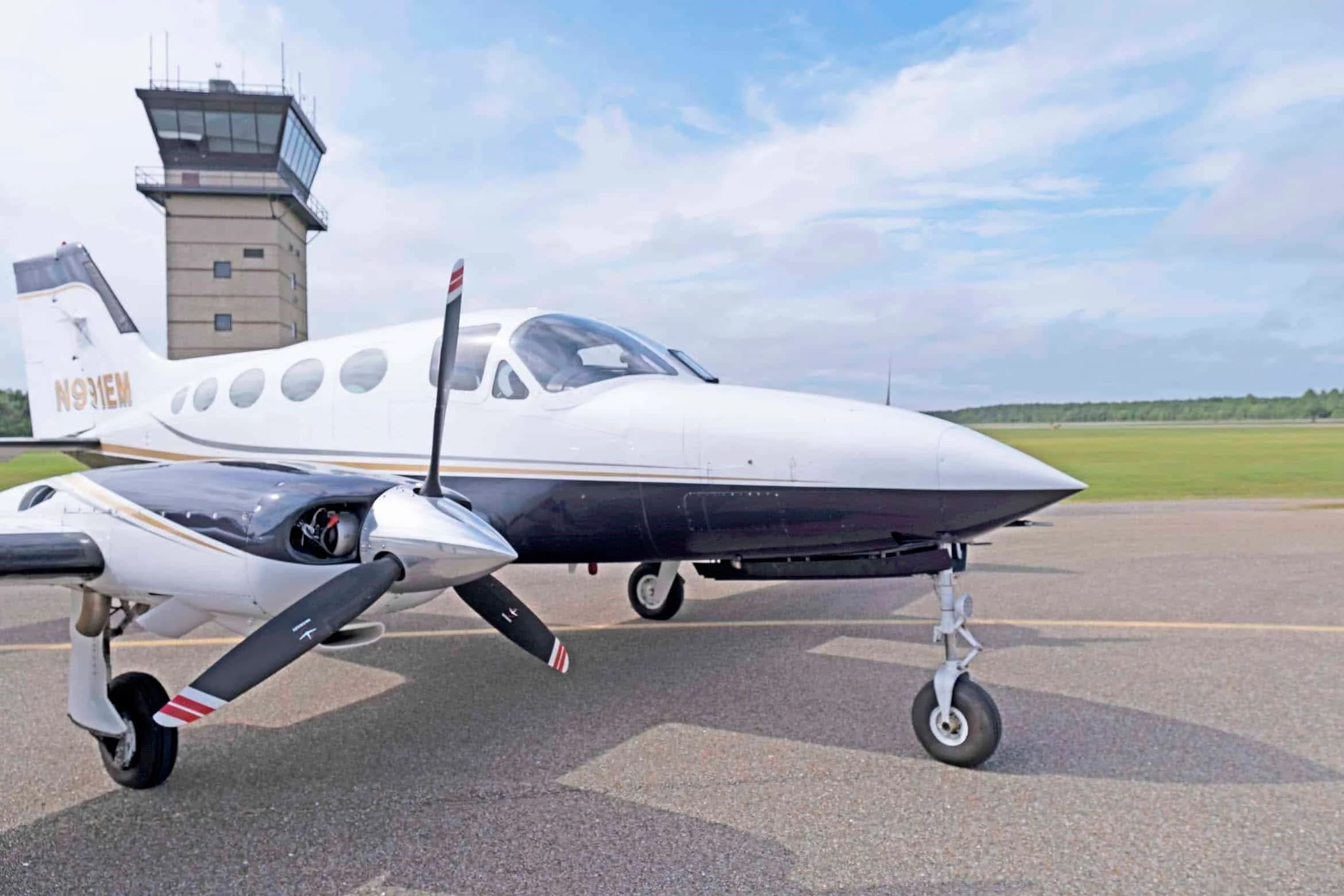 A front-quarter view of the white and dark blue Cessna 414 Chancellor, registration N991EM, with a control tower in the background.