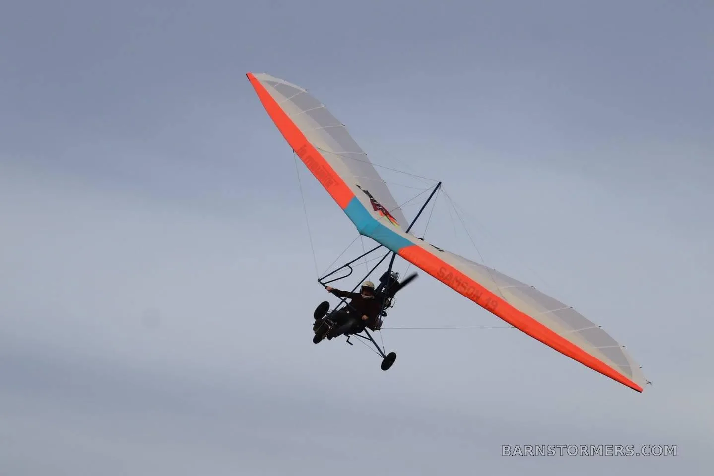 A powered hang glider with an orange, blue, and white wing, labeled 'SAMSON 19', flies against a clear sky.