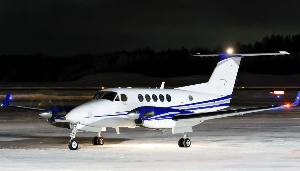 A white and blue Beechcraft King Air turboprop aircraft is parked on a snowy tarmac at night.