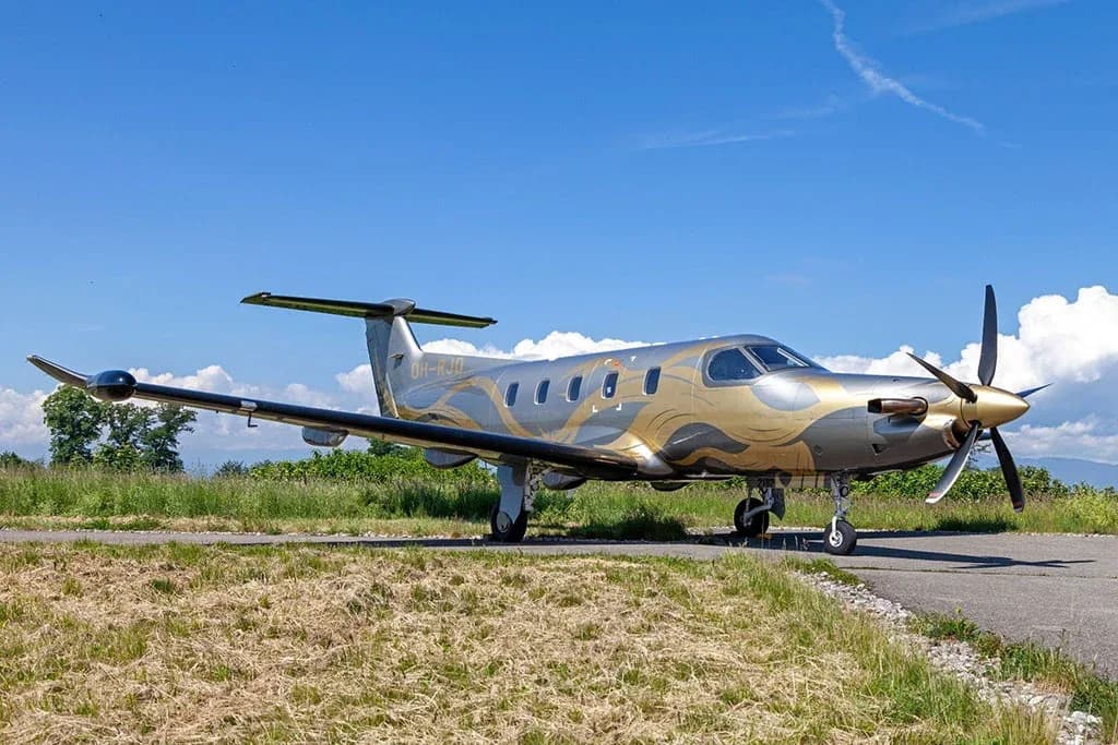 A silver and gold Pilatus PC-12 aircraft (OH-RJD) with a distinctive paint scheme, parked on an airfield with grass and a blue sky.