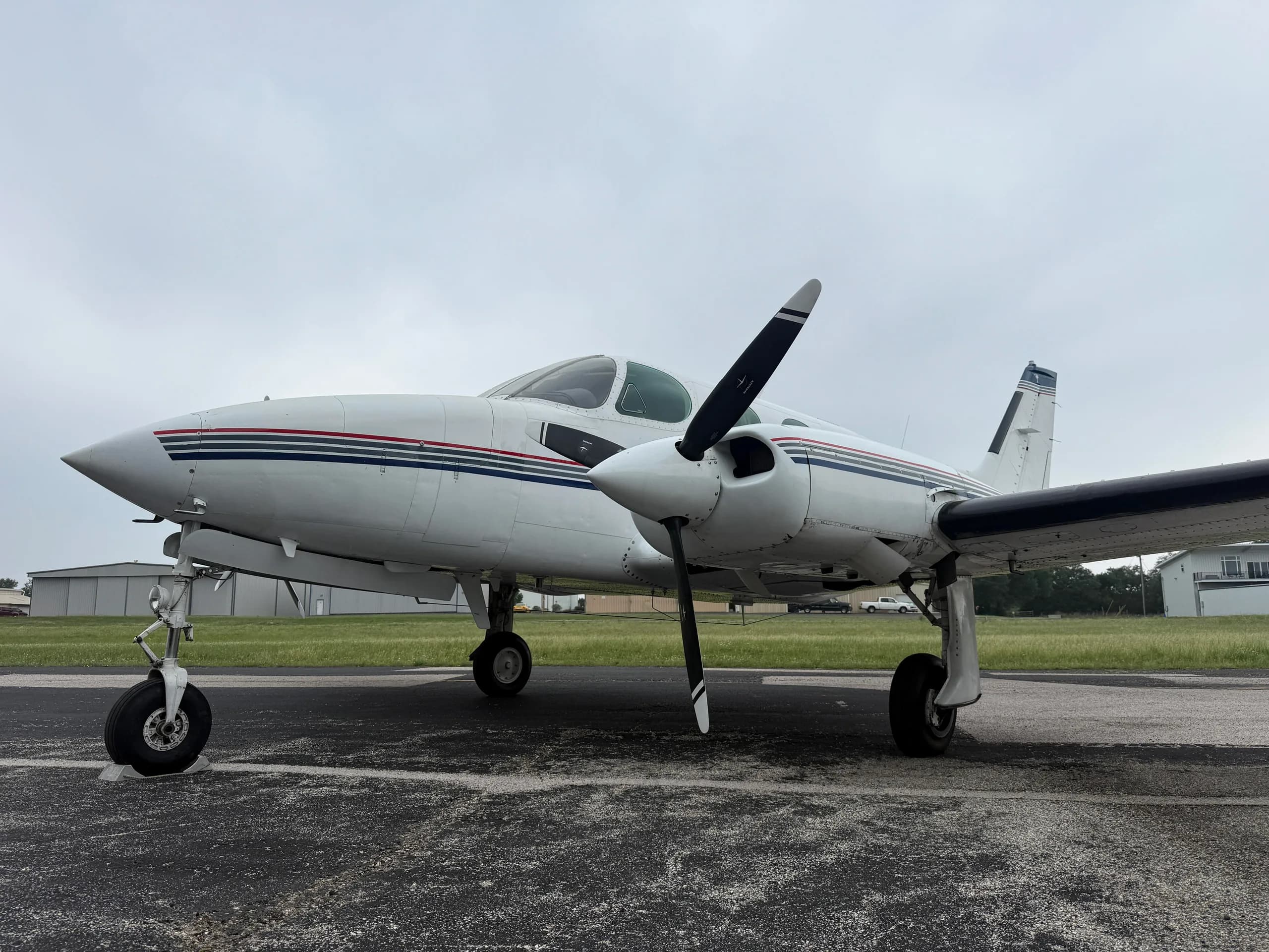 A white Cessna 335 aircraft with blue and red stripes on the fuselage and tail, seen from the front-left on an airfield.