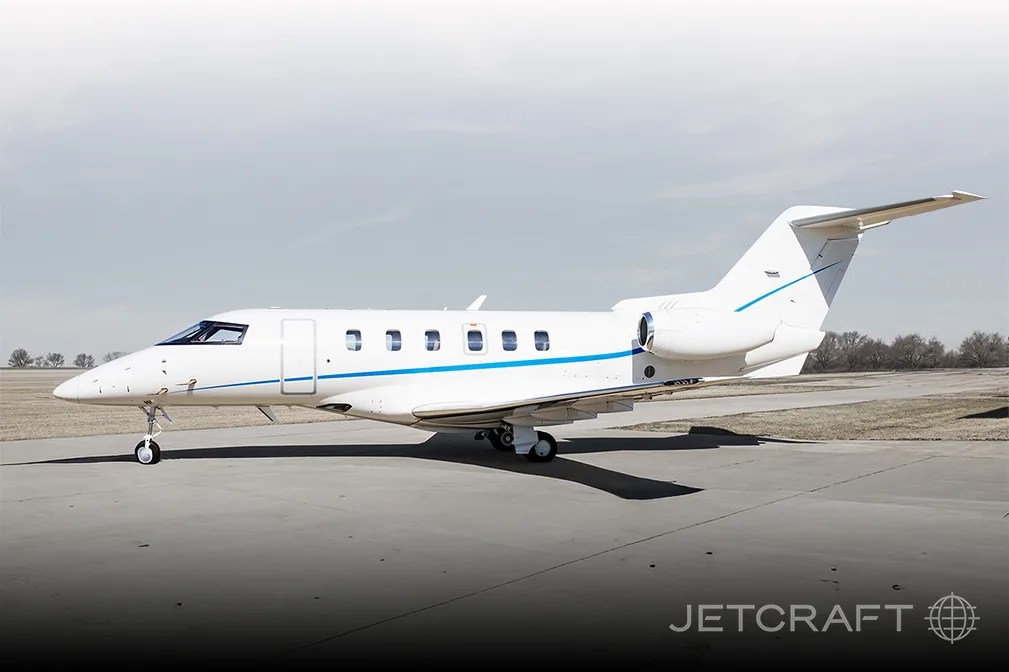 A white business jet with blue accent stripes is parked on an asphalt tarmac under a bright sky.