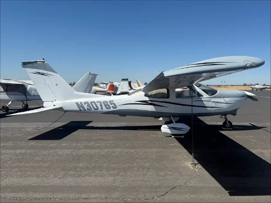 A white Cessna single-engine aircraft with grey and black stripes, registration N30765, parked on an airport tarmac on a sunny day.