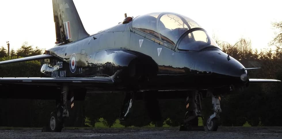 Side view of a black Royal Navy BAE Hawk jet aircraft parked on the ground at sunset.