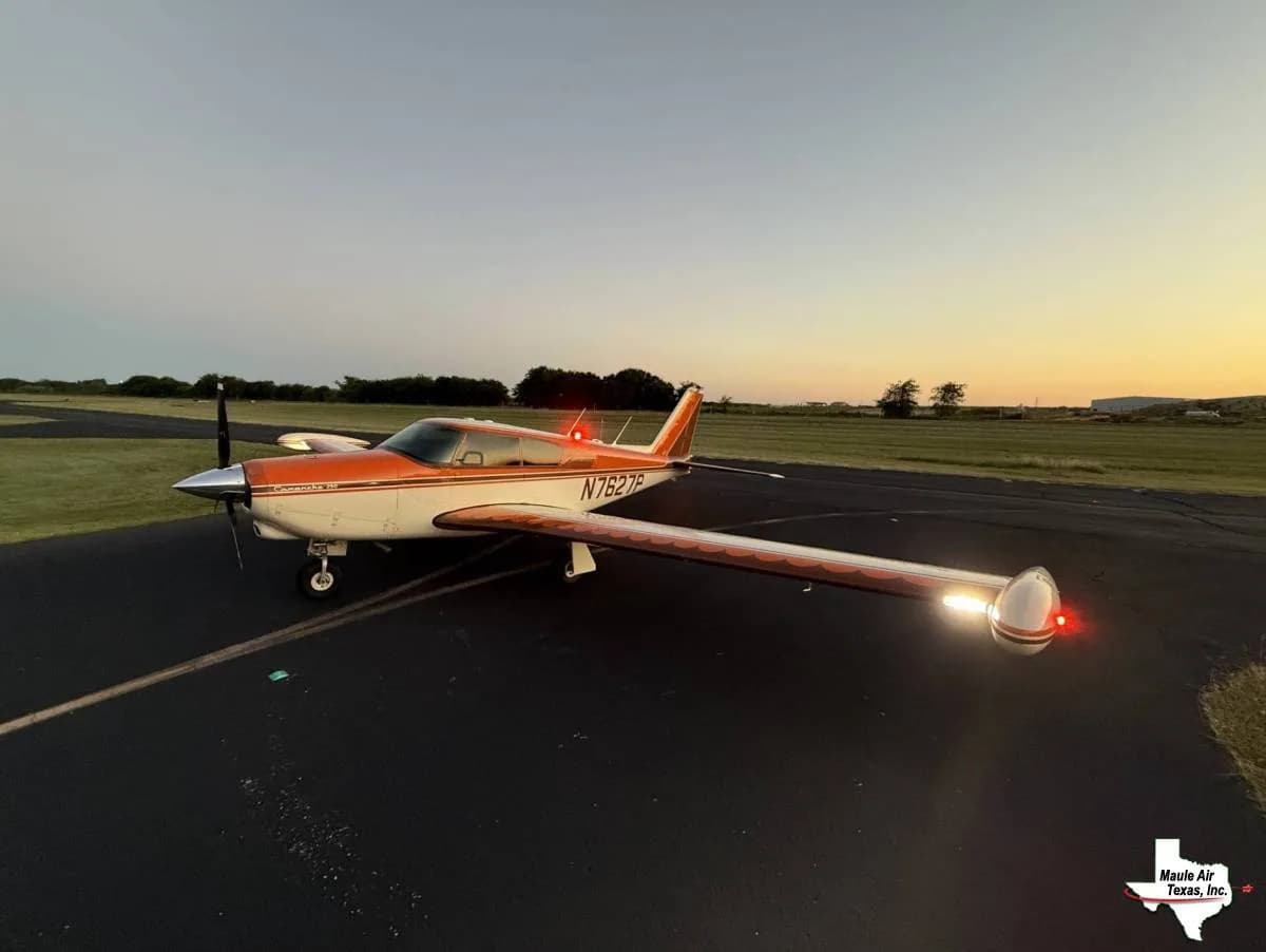 A Piper Comanche 250 aircraft, painted orange and white, with navigation lights illuminated, parked on an airfield at dusk.