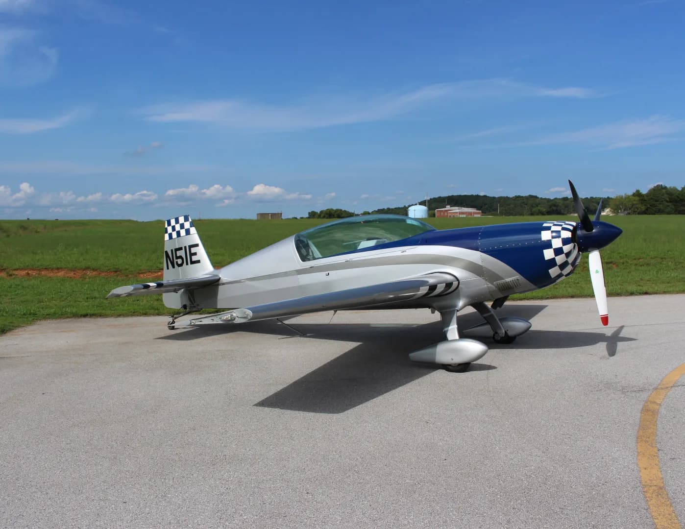 A silver and blue Extra EA-300/L aircraft with a checkered tail and nose cone parked on an asphalt runway.