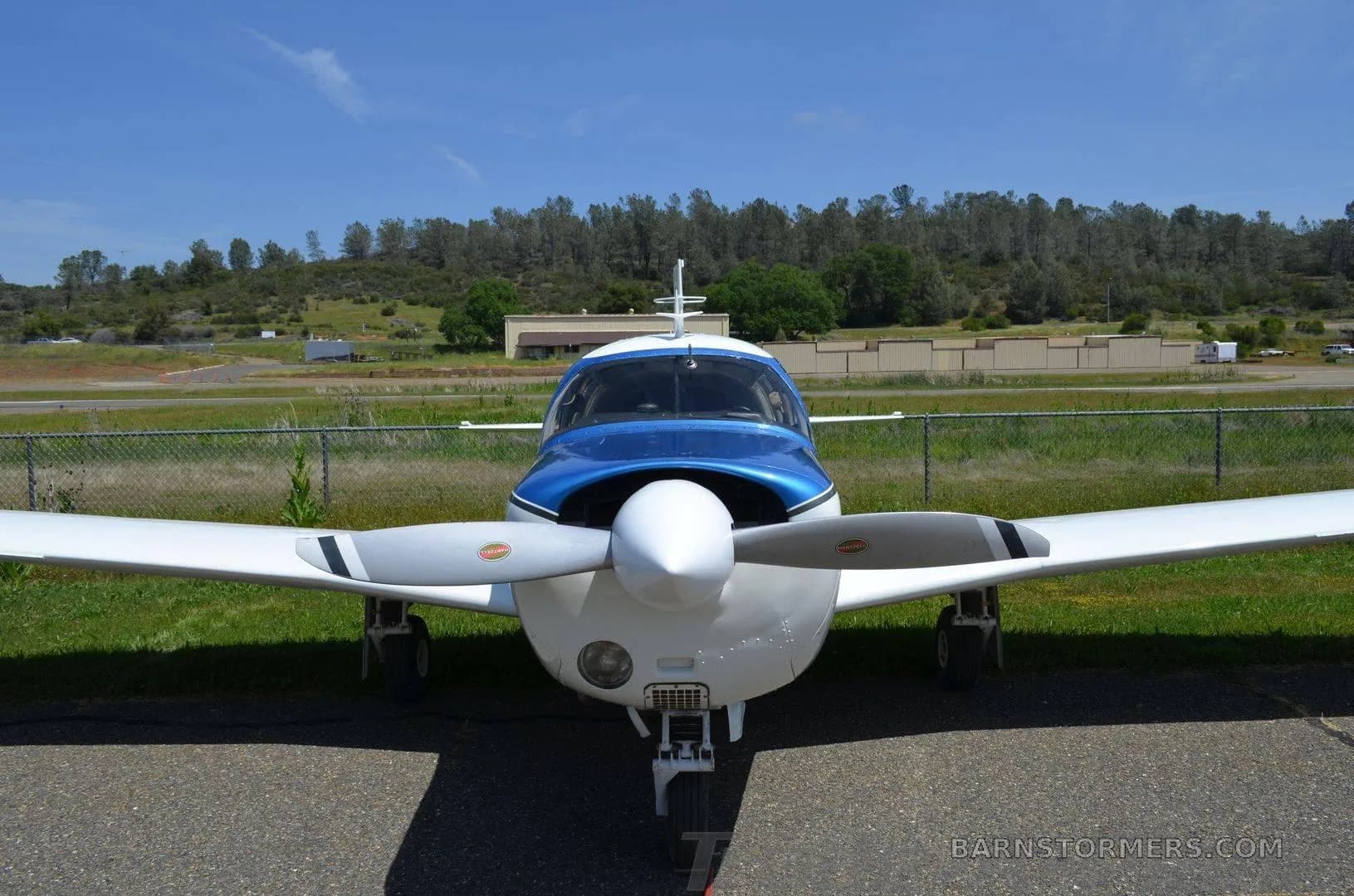 A white and blue single-engine propeller aircraft is parked head-on on an asphalt surface, with a grassy hill and trees in the background.