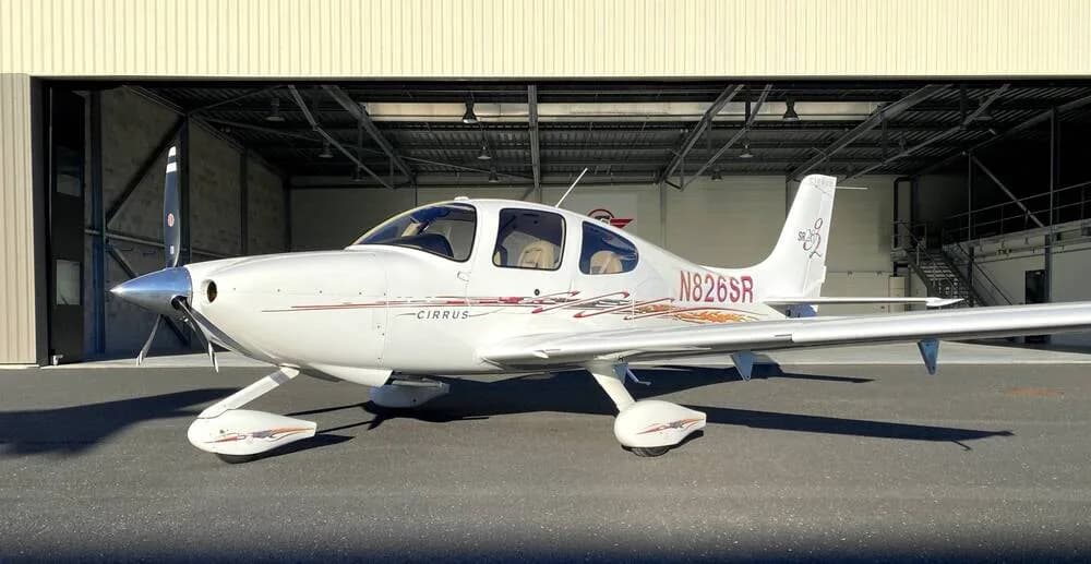 A three-quarter front view of a white Cirrus SR22 aircraft with red and gold accent stripes, registration N826SR, parked in front of an open hangar.
