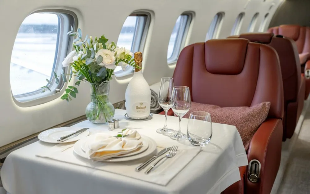 A set dining table with champagne, glasses, and flowers next to a window in a private jet cabin.