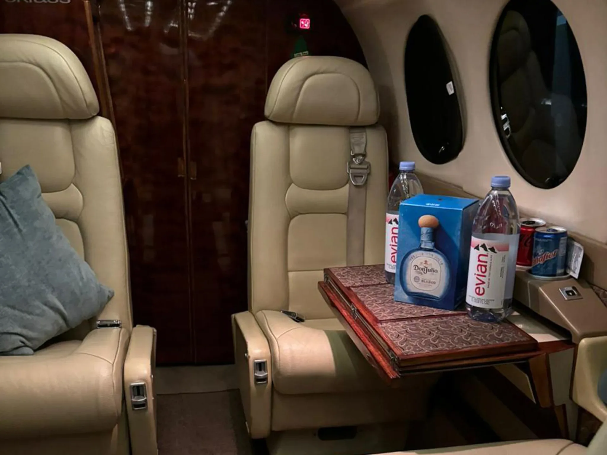 A close-up of two beige leather seats in an aircraft cabin with bottled water and a bottle of Don Julio tequila on a fold-out table.