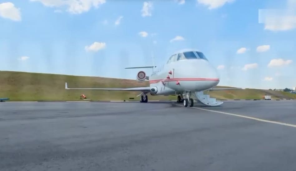 A white business jet with red stripes on the fuselage, parked on an tarmac with its main door open.
