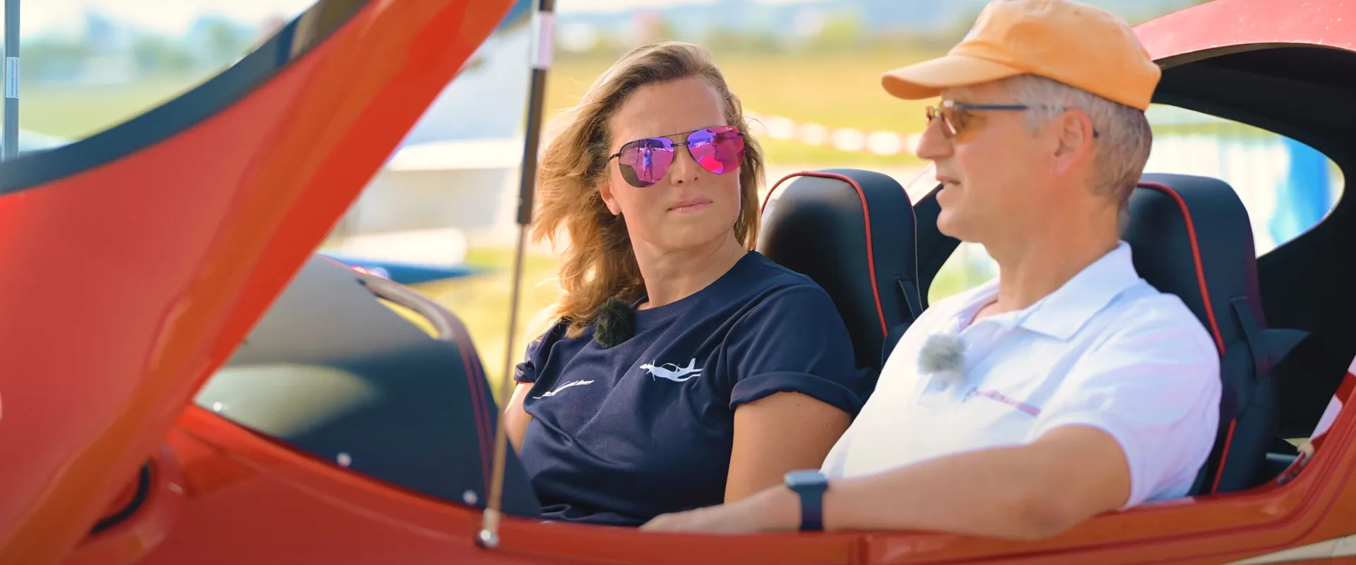 Two people conversing in the red cockpit of a light aircraft on an airfield.