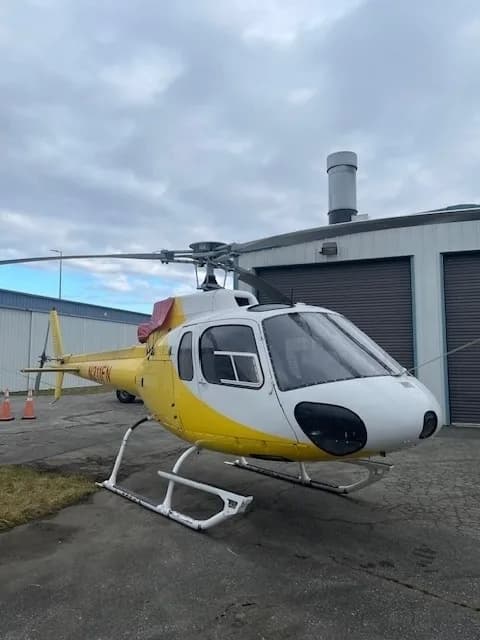 A white and yellow Eurocopter AS350 B3 helicopter, N211FN, is parked outdoors next to a hangar.