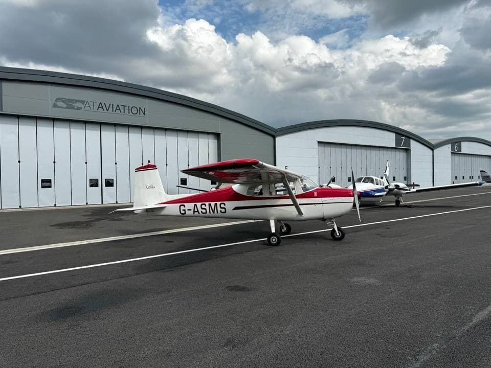 A white and red Cessna 150 G-ASMS parked on the tarmac with other aircraft and hangars under a cloudy sky.