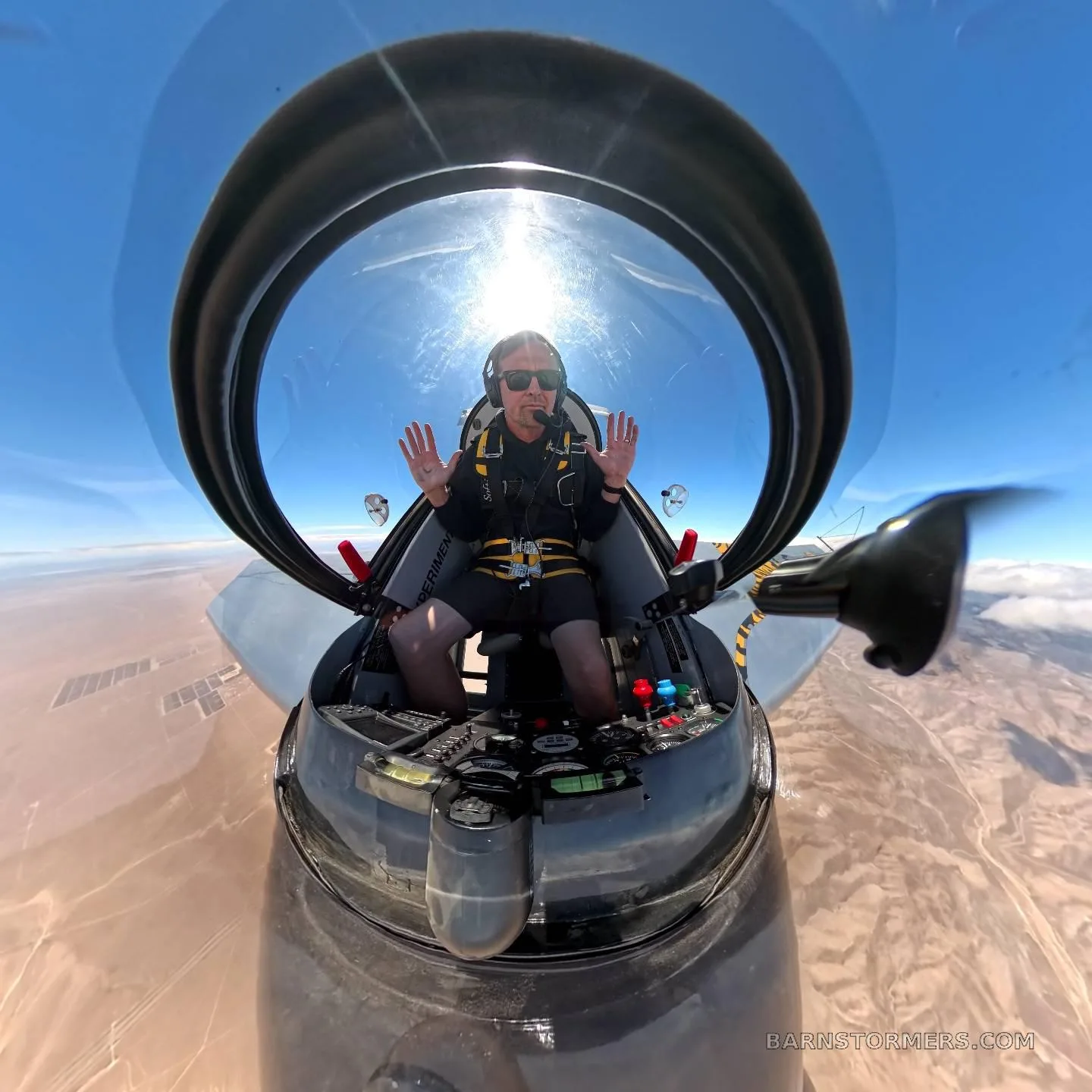 A pilot in a small aircraft with an open canopy raises both hands while flying over a desert landscape.