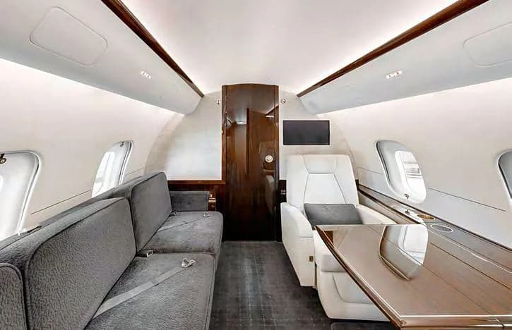 A section of the private jet interior with a grey sofa, a cream armchair, and a large wooden table.