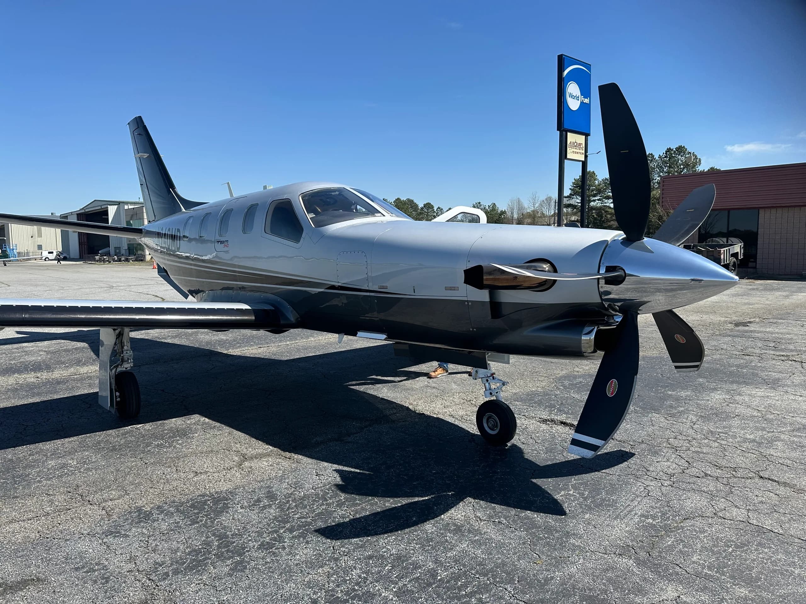 A silver and black TBM 940 aircraft with a large propeller, parked on an asphalt tarmac under a clear blue sky.