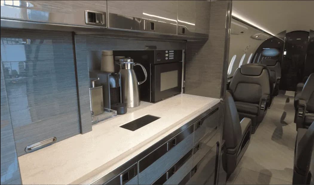 The galley area of a business jet cabin featuring a countertop, coffee maker, and microwave, with passenger seats visible in the background.