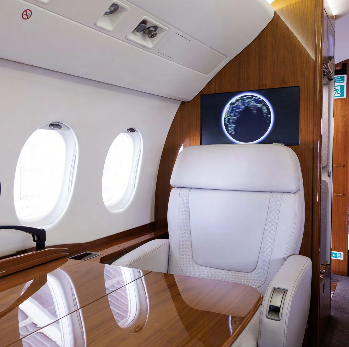 A single light grey leather seat in the business jet cabin with a polished wooden table and a screen displaying a globe.