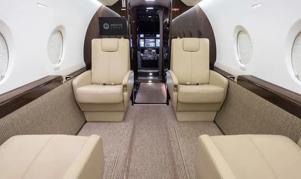 The luxurious interior cabin of the Gulfstream G550 features light-colored leather seats and dark wood accents, looking towards the cockpit.