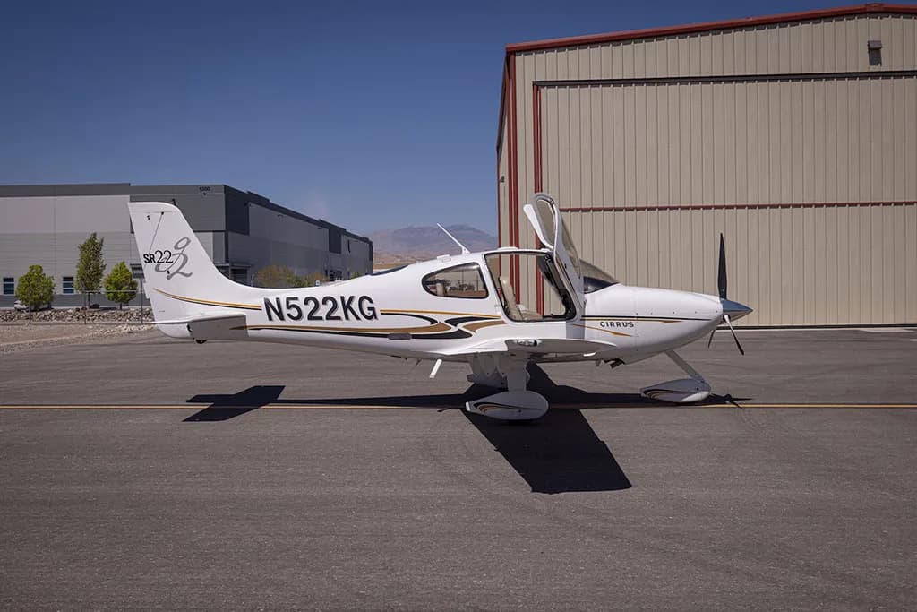 A side view of a white Cirrus SR22 aircraft with gold and black stripes, registration N522KG, parked on an airfield with its door open.