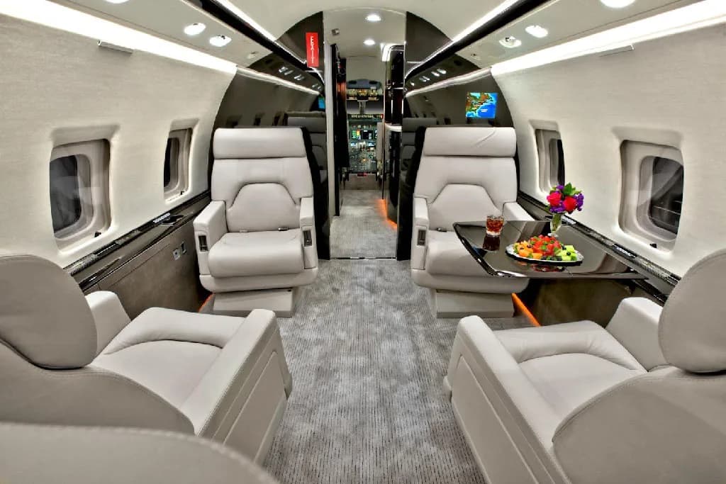 The luxurious interior cabin of a private jet, featuring light gray leather seats, a dark table with fruit, and a galley in the background.