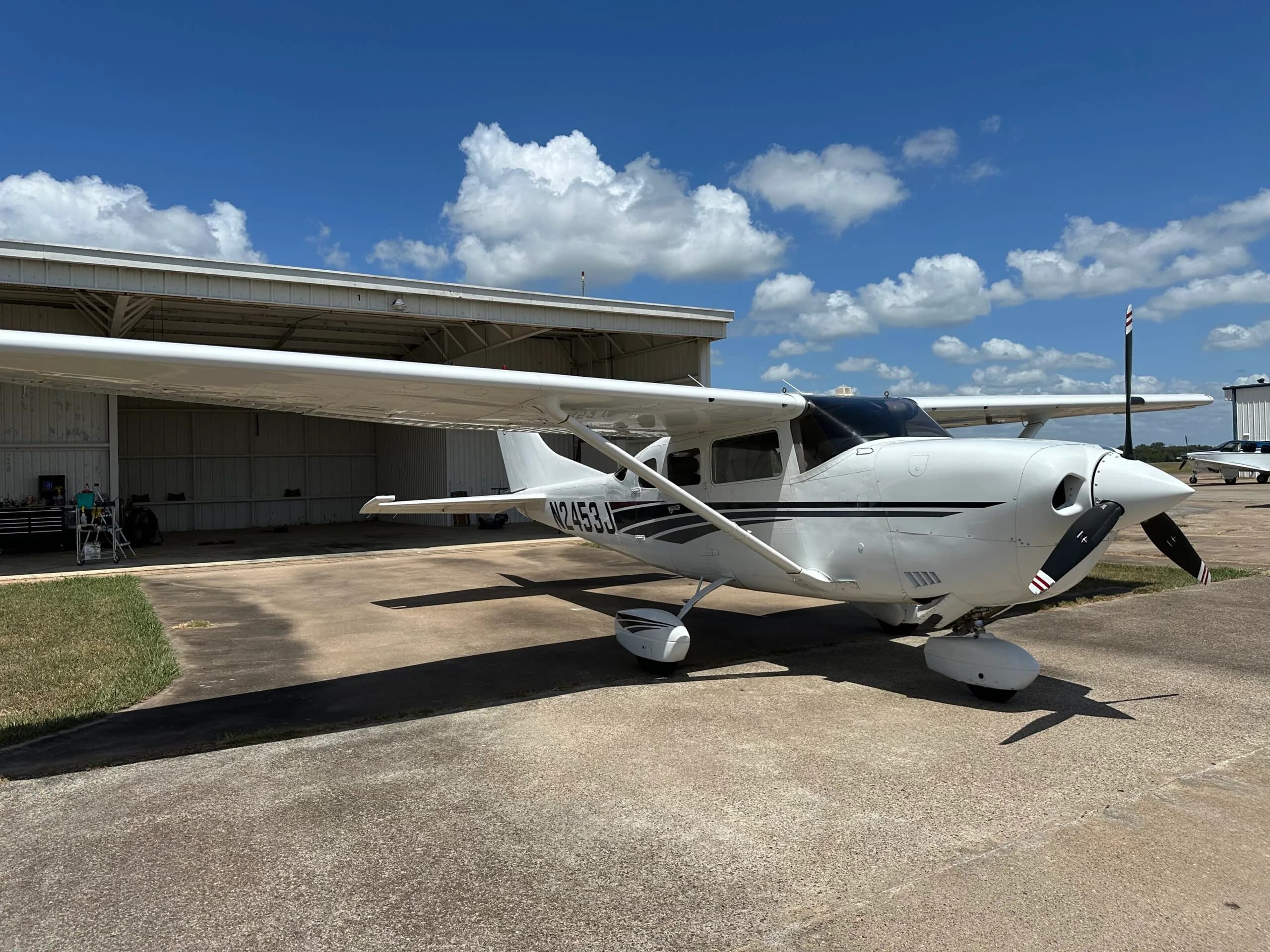 A white single-engine propeller aircraft with black and grey stripes, tail number N2453J, is parked on an airfield next to an open hangar under a blue sky.