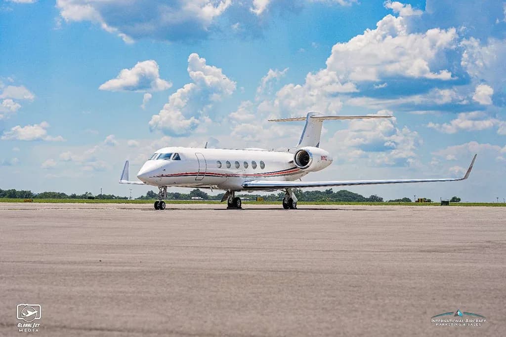 A side view of a white Gulfstream G550 aircraft with red and blue stripes, parked on an airfield under a blue sky.