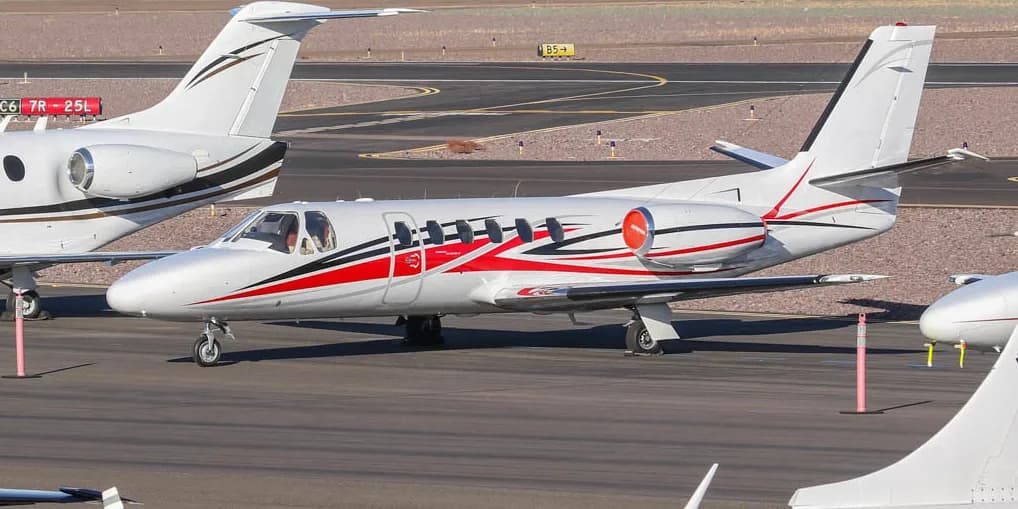 A white Cessna Citation business jet with red and black stripes parked on an airport tarmac.